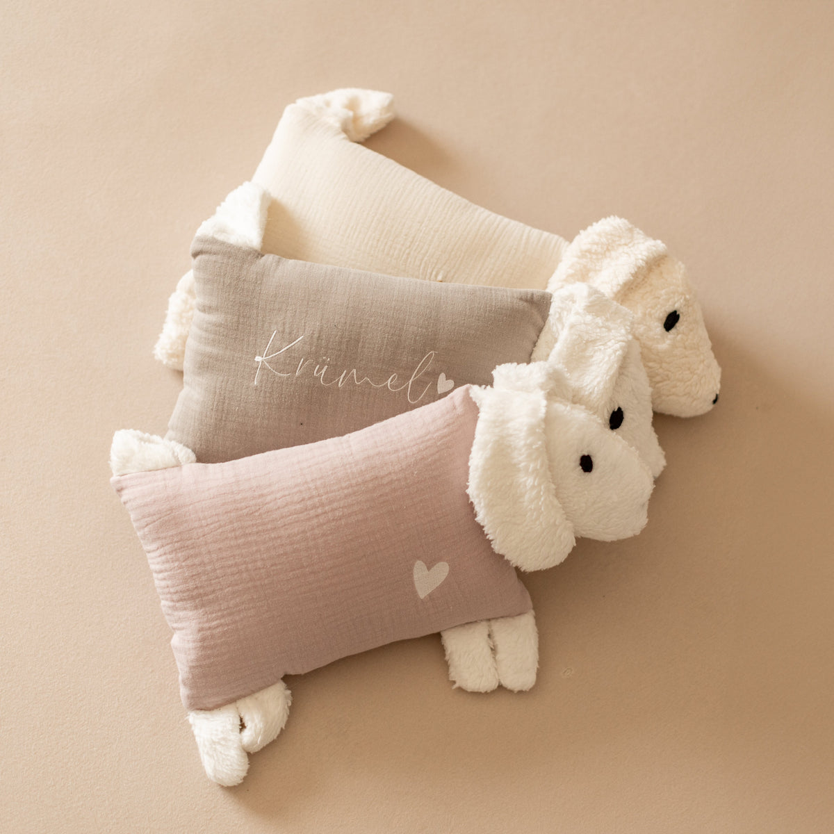 Drei Kuschelhunde mit naturfarbenem, beigem und rosa Stoffkörper liegen nebeneinander.